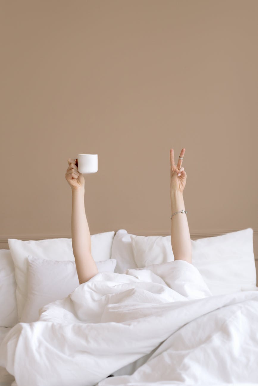 a person lying in bed holding up a cup of drink doing a peace sign